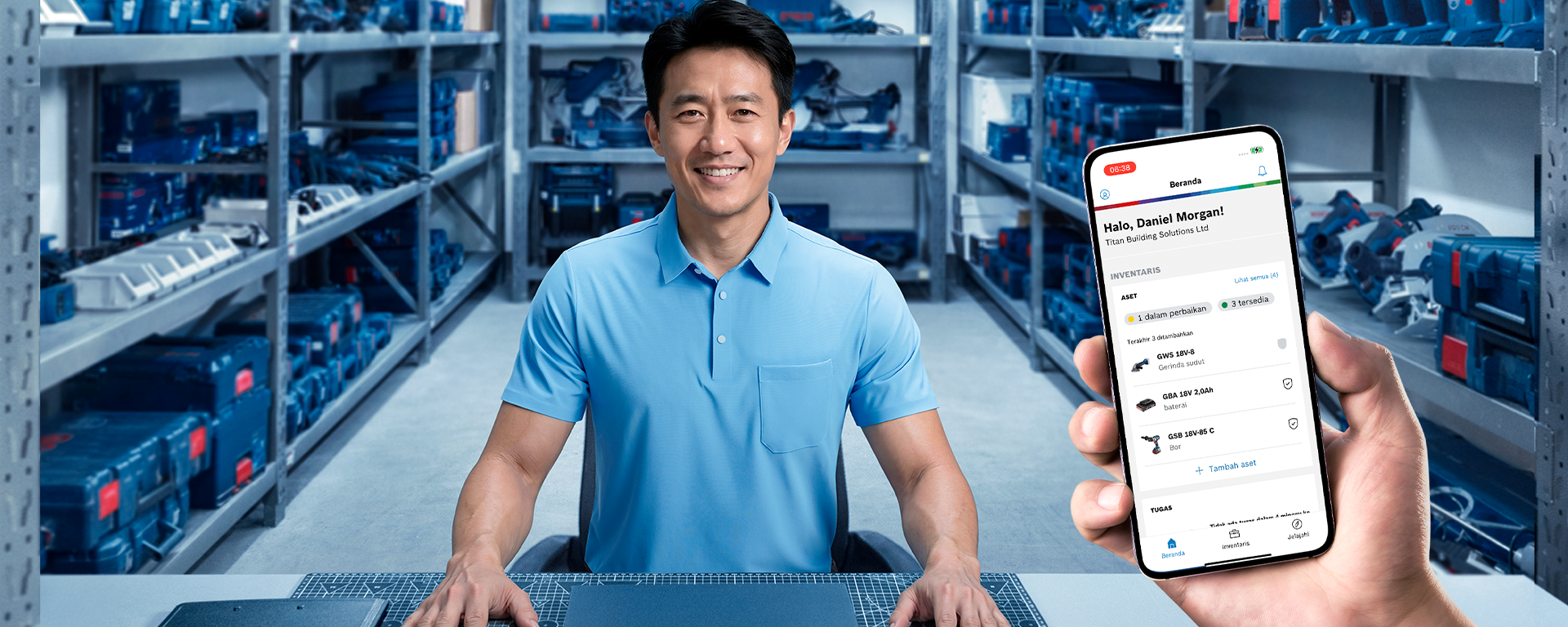 Seorang pria tersenyum mengenakan kaos polo biru duduk di meja, dikelilingi rak-rak penuh kotak alat.