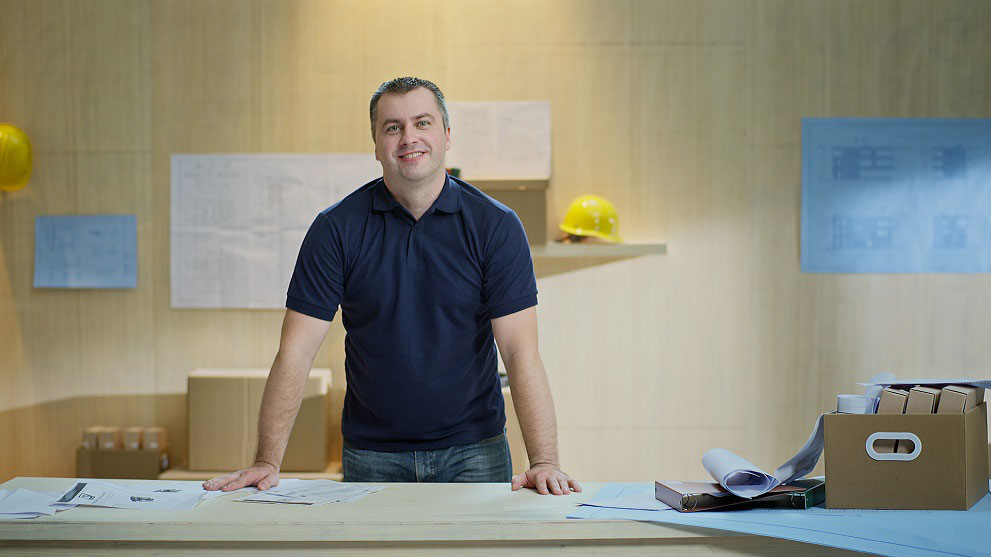 Seorang pria tersenyum dengan pakaian biru berdiri di meja dengan rencana bangunan dan folder. Helm kuning dan rencana terlihat di latar belakang.