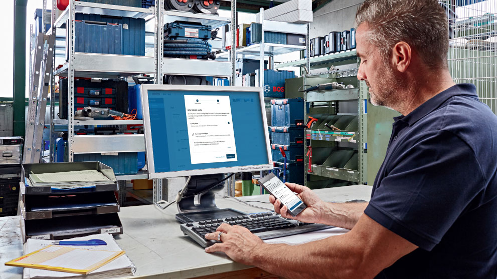 Seorang pria bekerja di komputer sambil memegang smartphone di tangannya. Di latar belakang terdapat rak dengan kotak alat dan bahan.