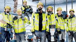 Enam pekerja konstruksi dengan helm kuning dan rompi peringatan berdiri di lokasi konstruksi.