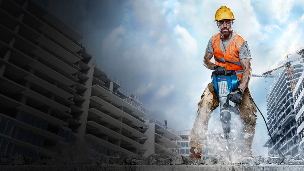 Seorang pria dengan helm pelindung dan rompi oranye sedang bekerja dengan palu Bosch.