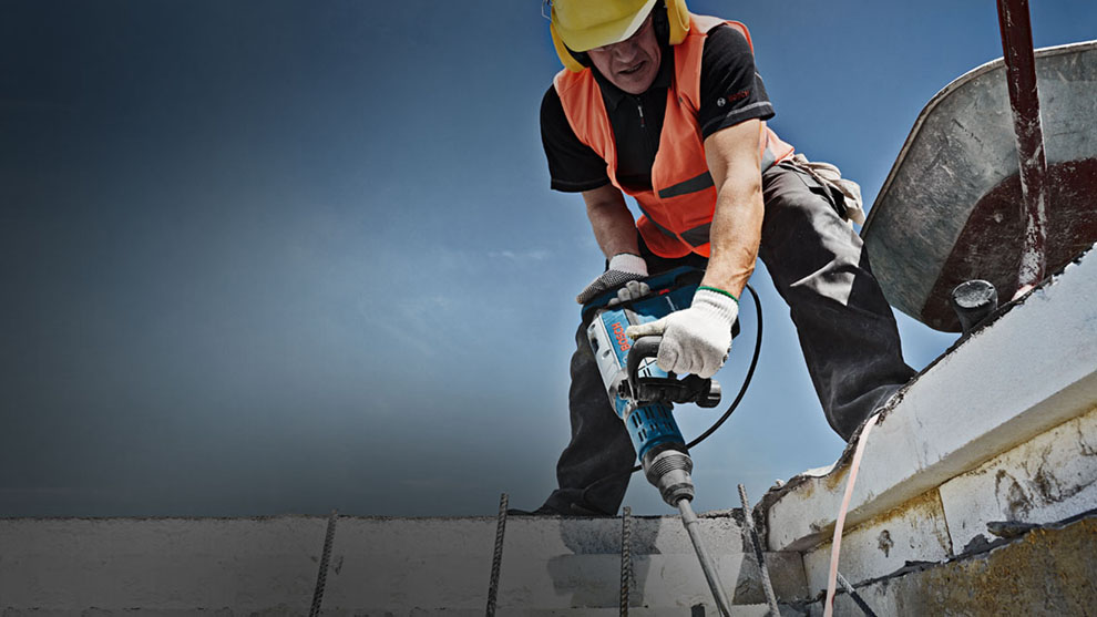 Pekerja dengan rompi keselamatan menggunakan bor Bosch di lokasi konstruksi.