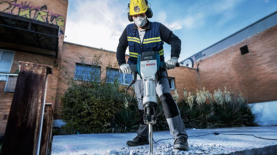 Seorang pekerja dengan palu Bosch sedang mengolah beton di luar ruangan.