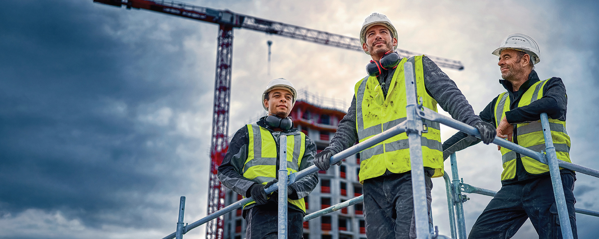 Tiga pekerja konstruksi mengenakan rompi keselamatan berdiri di atas perancah, melihat ke arah crane dan lokasi konstruksi.