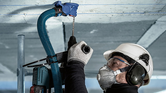 Seorang tukang yang mengenakan helm dan pelindung telinga sedang mengebor di langit-langit.