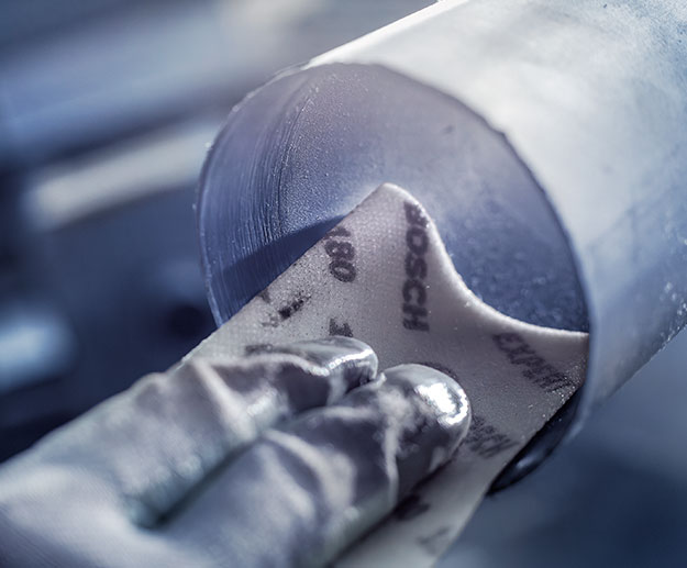 Sebuah tangan memegang kertas amplas Bosch pada sepotong logam bulat.